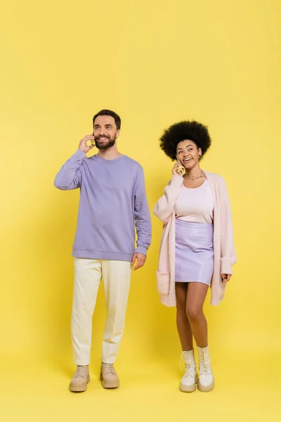 Full length of smiling interracial couple in stylish clothes talking on smartphones on yellow background — Photo de stock