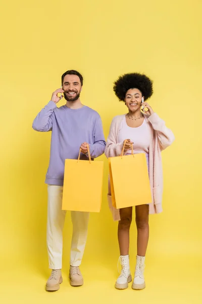 Full length of happy interracial couple holding shopping bags and talking on smartphones on yellow background — Stockfoto