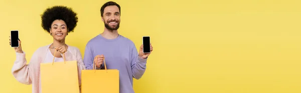 Cheerful bearded man and curly african american woman with shopping bags showing smartphones with blank screen isolated on yellow, banner — Stockfoto