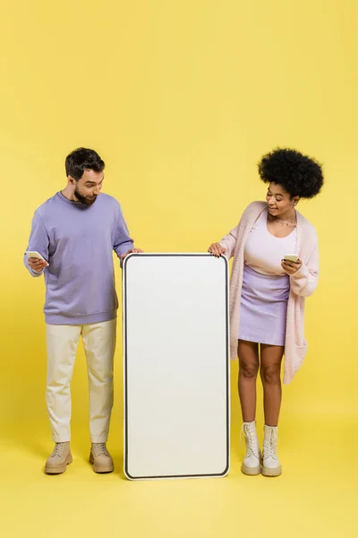 Full length of amazed interracial couple with gadgets looking at huge smartphone template on yellow background — Stock Photo