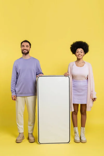 Полная длина радостной и стильной межрасовой пары, стоящей рядом с белым шаблоном мобильного телефона на желтом фоне — стоковое фото