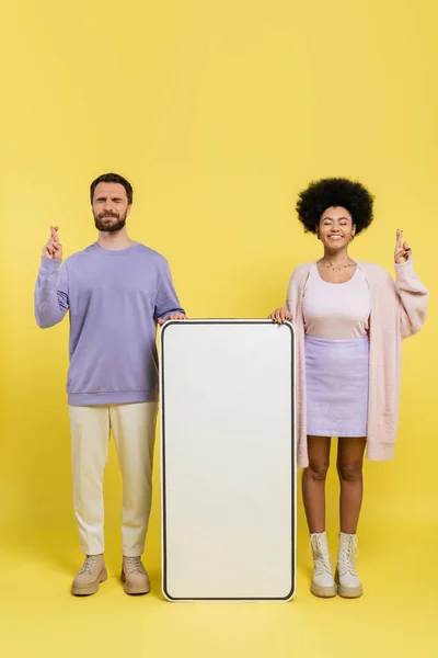 Повна довжина тривожного чоловіка і оптимістична афроамериканська жінка перетинає пальці біля макету смартфона на жовтому фоні — стокове фото