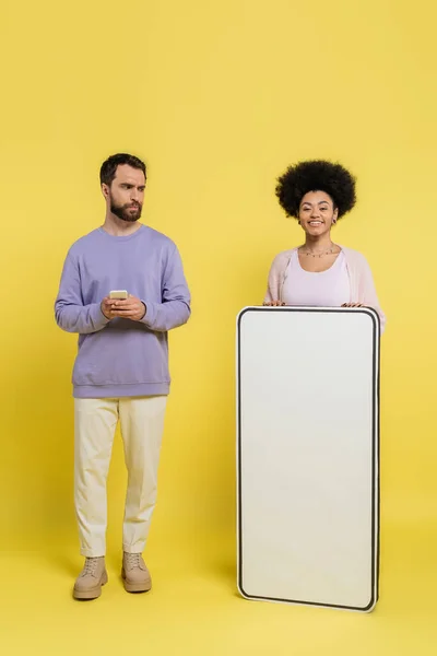 Bearded man with mobile phone looking at african american woman smiling near cardboard phone mock-up on yellow background - foto de stock