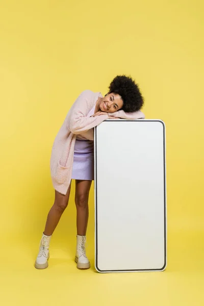 Full length of happy and trendy african american woman posing with huge template of smartphone on yellow background — Stock Photo