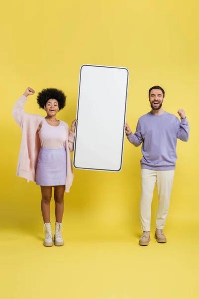 Full length of excited interracial couple rejoicing near carton mock-up of mobile phone on yellow background - foto de stock