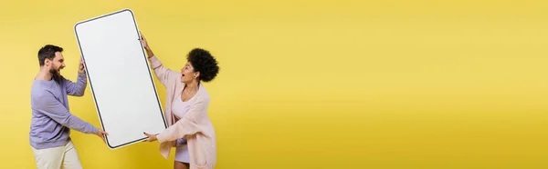 Astonished interracial couple holding big template of smartphone isolated on yellow, banner - foto de stock