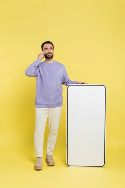 Full length of bearded man talking on smartphone near huge phone template on grey background — Stock Photo
