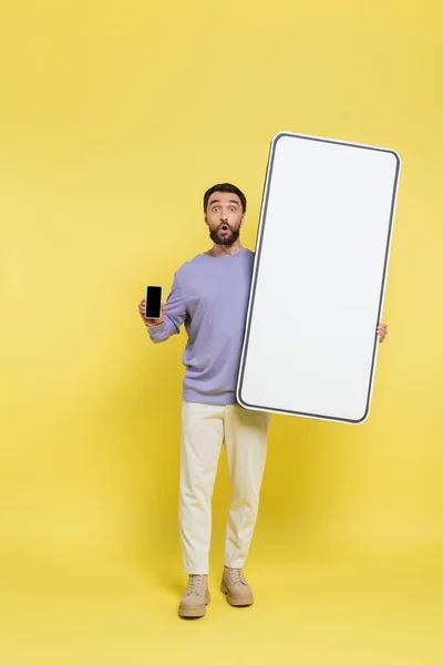 Full length of surprised man showing smartphone with blank screen near white template of mobile phone on grey background — Photo de stock