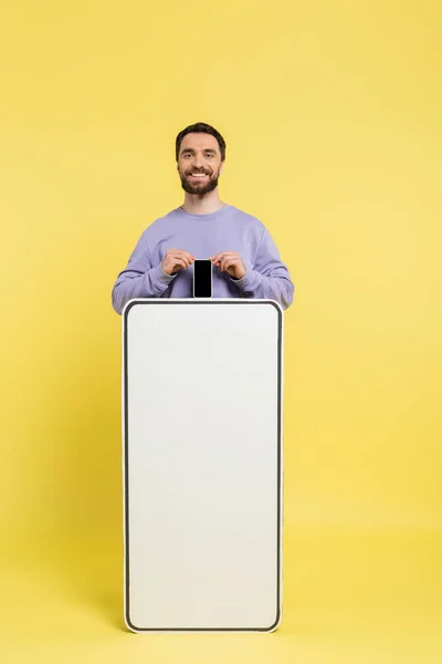 Bearded man smiling at camera and showing smartphone with blank screen near white phone template on yellow background — Stock Photo