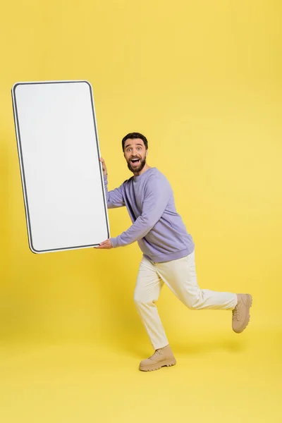 Full length of amazed bearded man looking at camera while running with template of cellphone on grey background — Foto stock