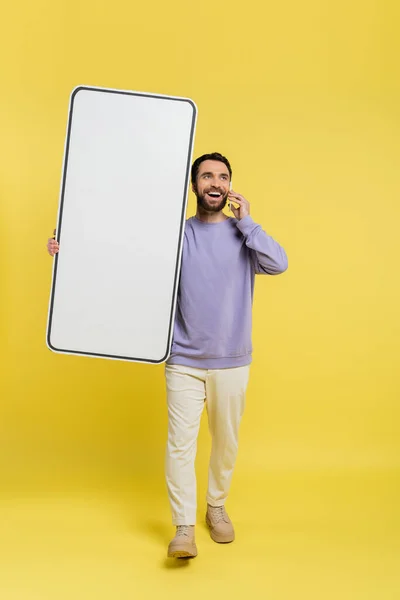 Full length of happy man walking with huge phone mock-up and talking on cellphone on grey background — Stock Photo