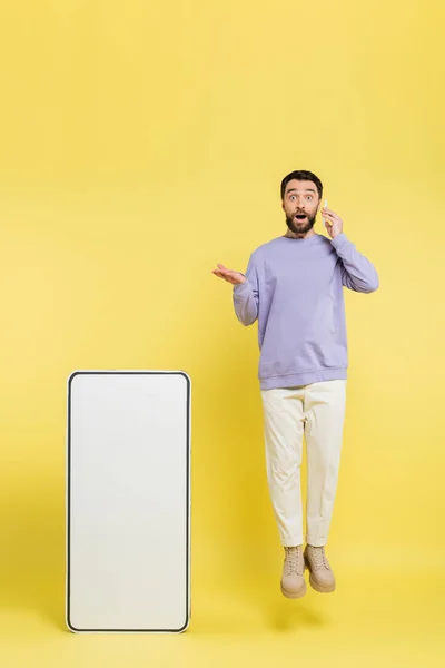 Amazed man talking on cellphone while levitating and pointing at huge mock-up of mobile phone on grey background — Stock Photo