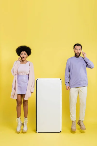 Full length of astonished interracial couple talking on smartphones near carton template of mobile phone on yellow background - foto de stock