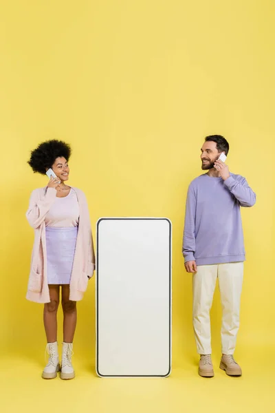 Full length of interracial couple talking on cellphone and smiling at each other near blank mock-up of smartphone on yellow - foto de stock