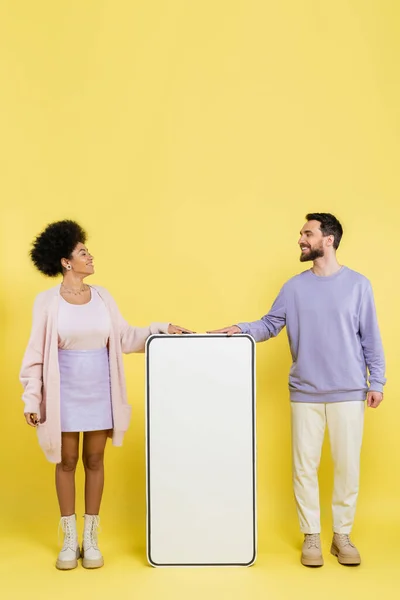 Full length on trendy interracial couple smiling at each other near white mock-up of mobile phone on yellow background - foto de stock