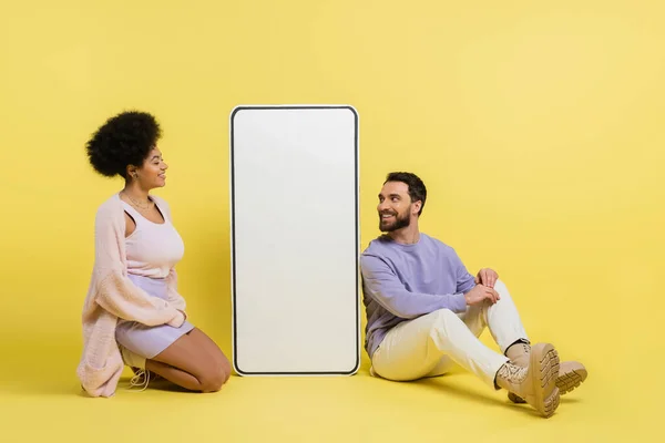 Полная длина на веселой и модной межрасовой пары, сидящей рядом с белым шаблоном смартфона на желтом фоне — стоковое фото
