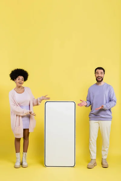 Full length of pleased multiethnic couple looking at camera and pointing at huge mobile phone template on yellow background — Stockfoto