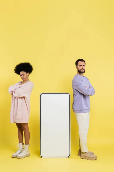 Full length of displeased interracial couple standing with crossed arms near huge phone template on yellow background - foto de stock