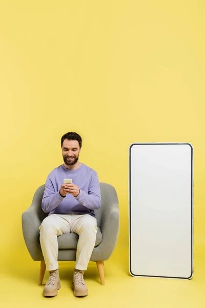 Full length of pleased man using smartphone while sitting in armchair near huge template of mobile phone on grey background — Stock Photo