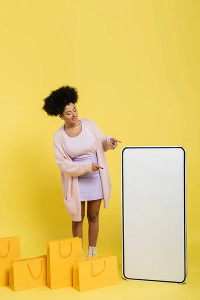 Trendy african american woman pointing at huge template of mobile phone near shopping bags on yellow background — Stock Photo