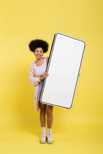 Full length of cheerful african american woman in leather boots holding huge template of smartphone on yellow background - foto de stock