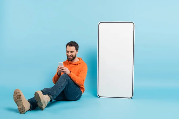 Full length of smiling bearded man messaging on smartphone while sitting near huge phone template on blue background — Stock Photo
