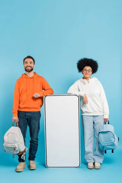 Full length of interracial students with backpacks smiling at camera near huge phone template on blue background — Stock Photo