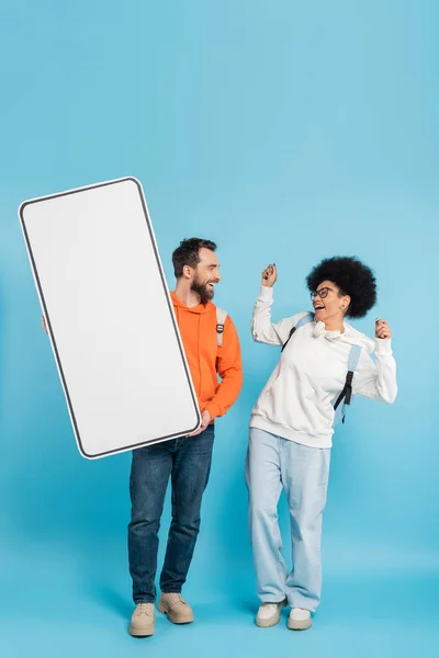 African american woman laughing near bearded student holding huge template of smartphone on blue background — Fotografia de Stock
