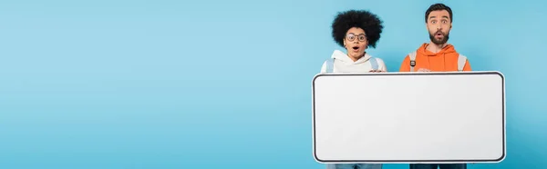 Amazed interracial students looking at camera near huge phone mock-up isolated on blue, banner — Stock Photo