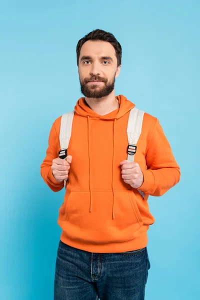 Bearded brunette student in orange hoodie holding backpack and looking at camera isolated on blue — Stockfoto