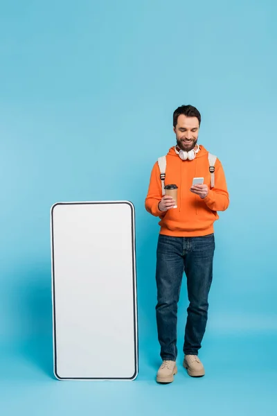 Full length of smiling student with coffee to go using smartphone near carton mock-up of cellphone on blue background — Fotografia de Stock