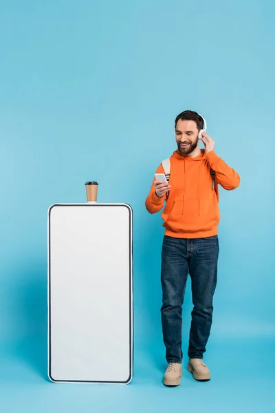 Full length of bearded student in wireless headphones standing with smartphone near phone mock-up and paper cup on blue background — Stock Photo