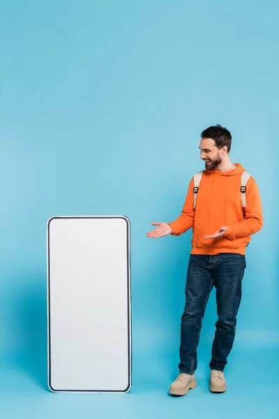 Full length of happy bearded student pointing at big template of mobile phone on blue background — Stock Photo