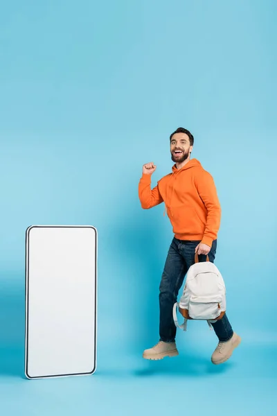 Full length of cheerful bearded student with backpack showing win gesture while levitating near phone template on blue background — Stockfoto