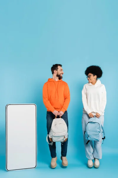 Full length of happy multiethnic students with backpacks looking at each other near smartphone template on blue background — Stockfoto