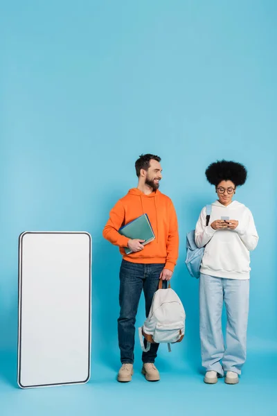 African american woman using smartphone near bearded student and white phone template on blue background — Photo de stock