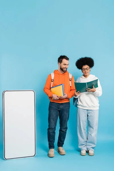 Full length of african american woman looking in copybook near smiling student and carton smartphone template on blue background — Fotografia de Stock