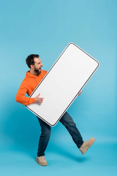 Full length of excited man holding huge template of smartphone on blue background — Stock Photo