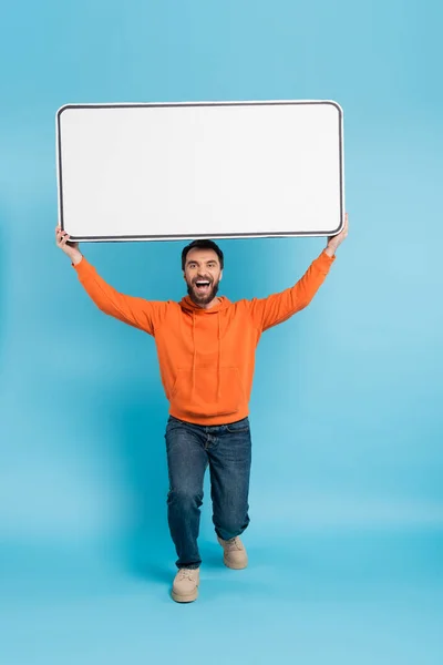 Full length of excited man in orange hoodie and jeans holding big phone template above head on blue background - foto de stock