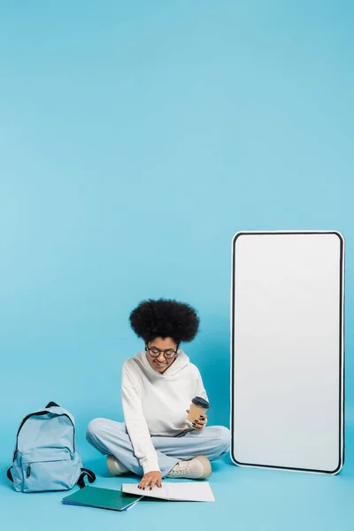 Афроамериканець з паперовою чашею дивиться в зошит, сидячи біля рюкзака і макетом з білого телефону на синьому — стокове фото