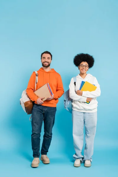 Full length of trendy interracial couple of students standing with copybooks and backpacks on blue — Stock Photo
