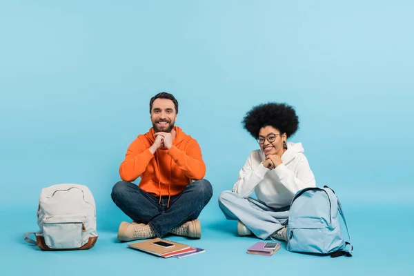 Glückliche multiethnische Studenten, die mit überkreuzten Beinen neben Rucksäcken und Notizbüchern mit Smartphones auf blau sitzen — Stockfoto