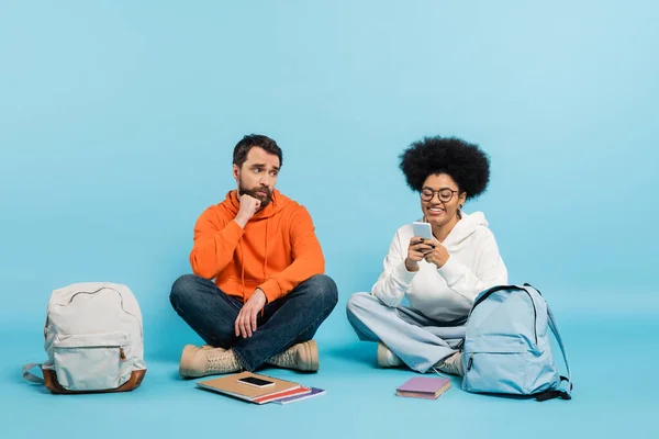 Cheerful african american student using smartphone near offended boyfriend sitting on blue background — Stock Photo