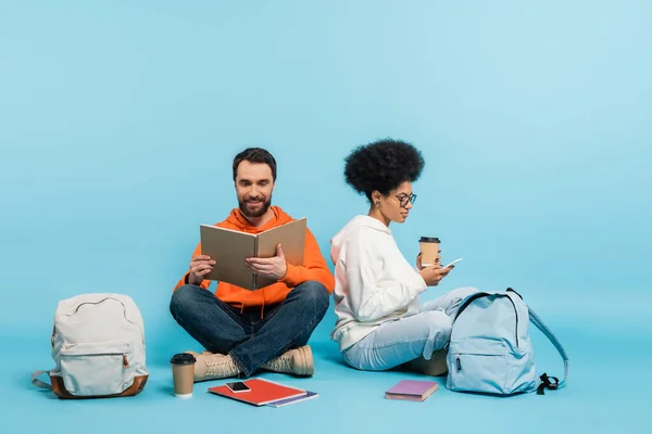 Bearded student looking in notebook near african american girlfriend sitting with smartphone on blue background - foto de stock