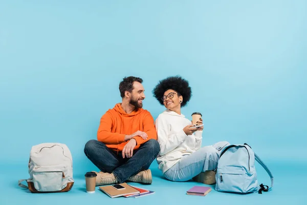 Happy multiethnic students sitting near backpacks and copybooks and smiling at each other on blue background — Stockfoto