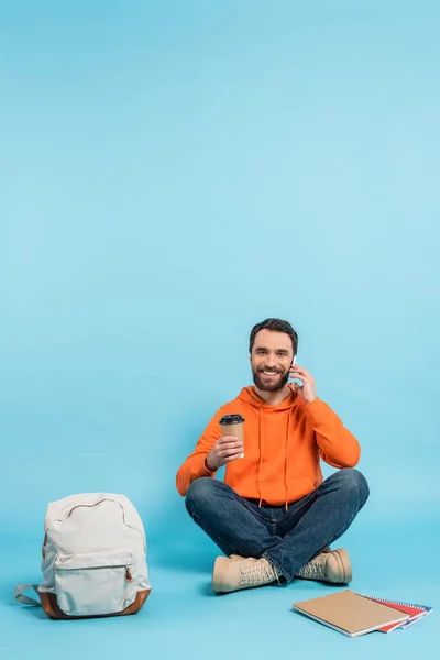 Lächelnder Student, der mit Coffee to go in der Nähe des Rucksacks sitzt und mit dem Smartphone spricht — Stockfoto