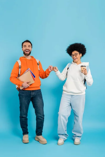 Full length of multiethnic students with notebooks and takeaway drink doing fist bump while smiling at camera on blue background — Stockfoto