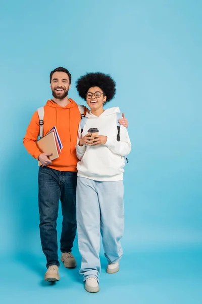 Bearded student with notebooks hugging african american girlfriend with paper cup while walking on blue - foto de stock