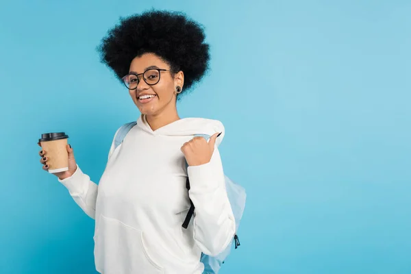 Cheerful african american student in eyeglasses holding takeaway drink and pointing with thumb isolated on blue — Stock Photo
