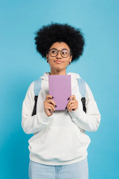 Étudiant afro-américain rêveur et souriant avec sac à dos tenant le livre et regardant loin isolé sur bleu — Photo de stock